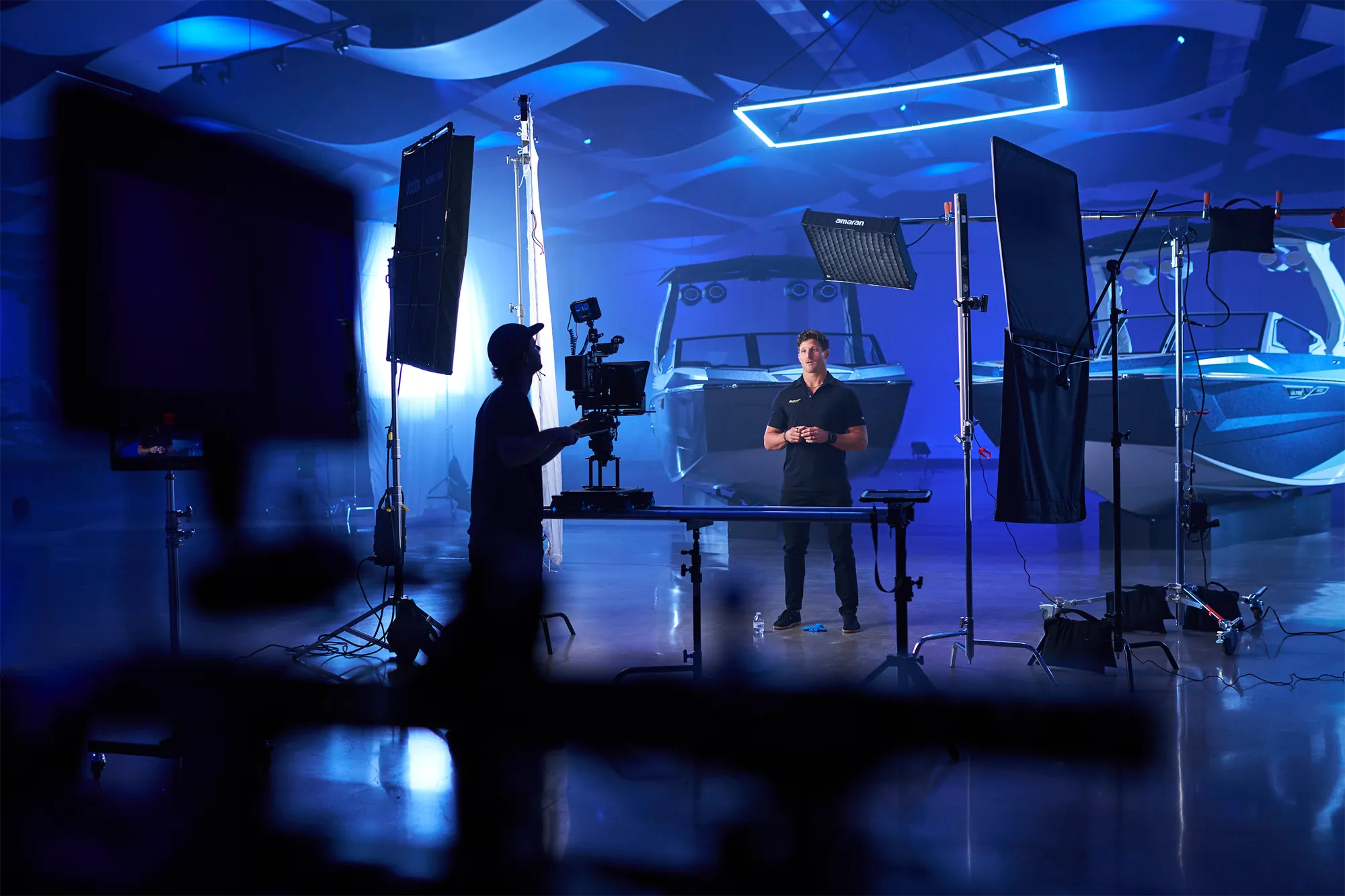 Joey Arcisz in a cap at a cinema camera on a slider, silhouetted against a blue-lit studio, a presenter in a black polo standing between two wake boats behind him, a rectangular LED tube overhead, and flags, a book light and a soft panel around the set.