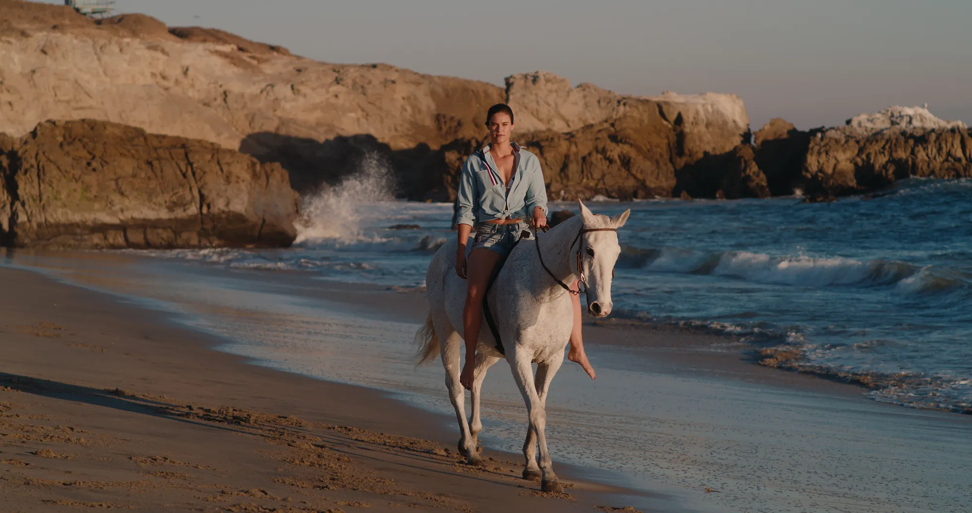 A woman in a pale blue shirt and denim shorts rides a white horse along the wet sand at the water's edge, a wave breaking against dark rocks behind her and a rocky point beyond, in low evening light.