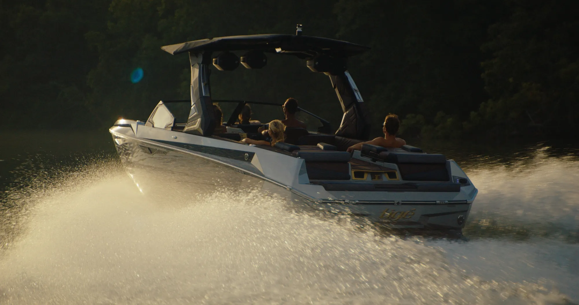 A wake boat with six people aboard, seen from behind and to one side, throws a wall of spray lit gold by low sun, a dark treeline behind.