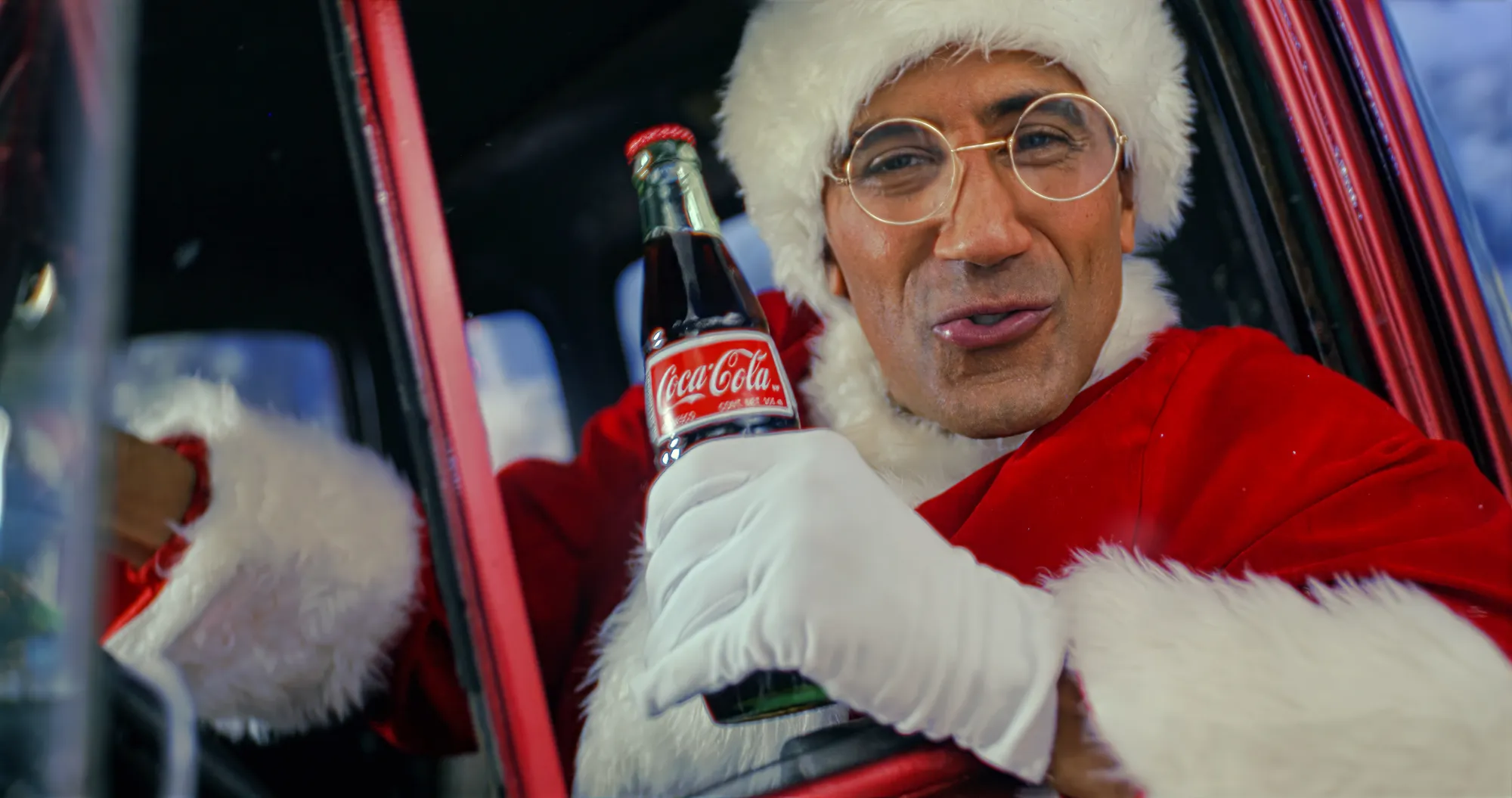 A performer in a Santa suit, round glasses and white gloves leans out of the window of a red vintage truck, holding a glass bottle of Coca-Cola up to the lens mid-line, snow falling behind.