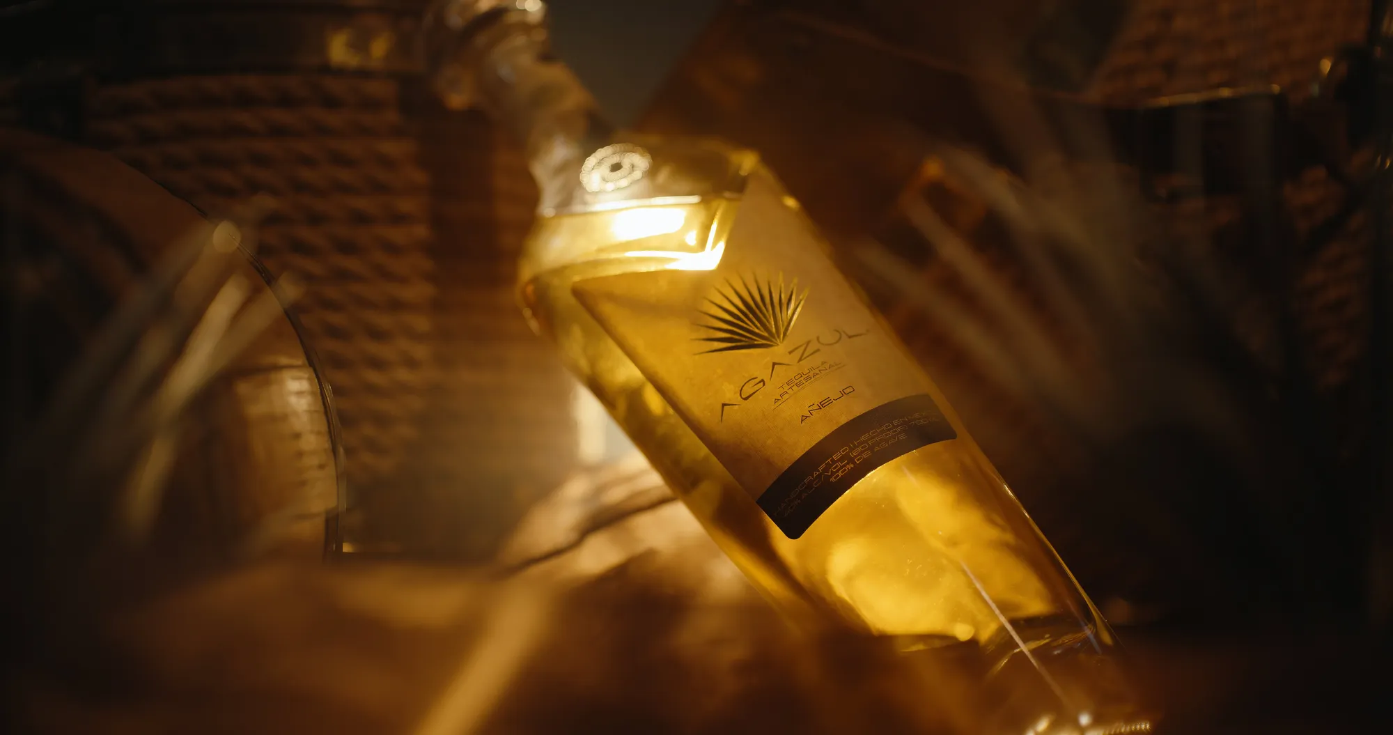 An Agazul añejo bottle lying at an angle in a woven basket, backlit so the amber tequila glows through the glass, agave leaves and grass softening around it in warm low light.