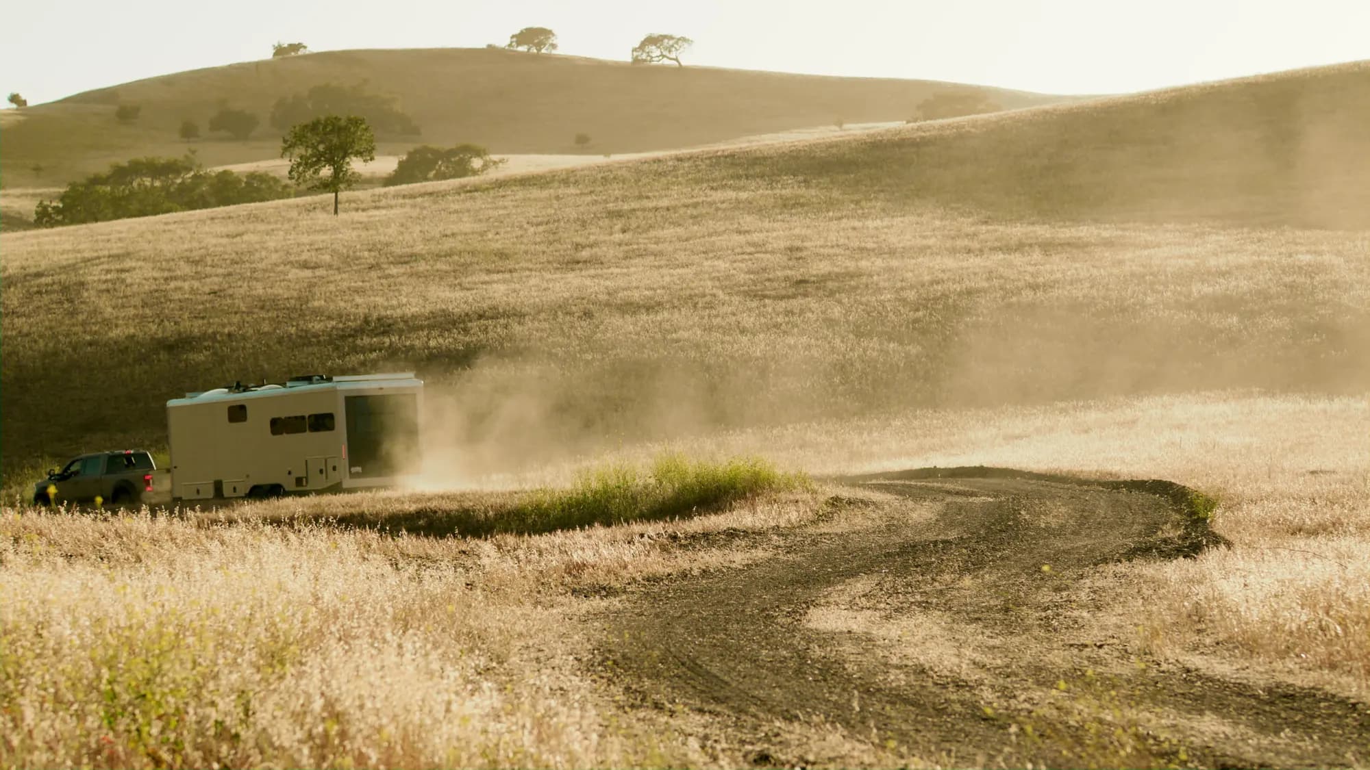 A white travel trailer behind a pickup climbs a dirt track through dry golden hills, trailing a long plume of dust, oaks along the ridge above, in hazy backlight.