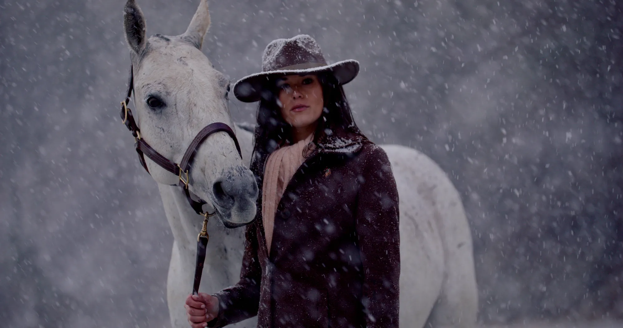A woman in a snow-dusted felt hat and dark coat stands beside a white horse with a hand on its bridle, snow falling heavily, trees fading into the storm behind.