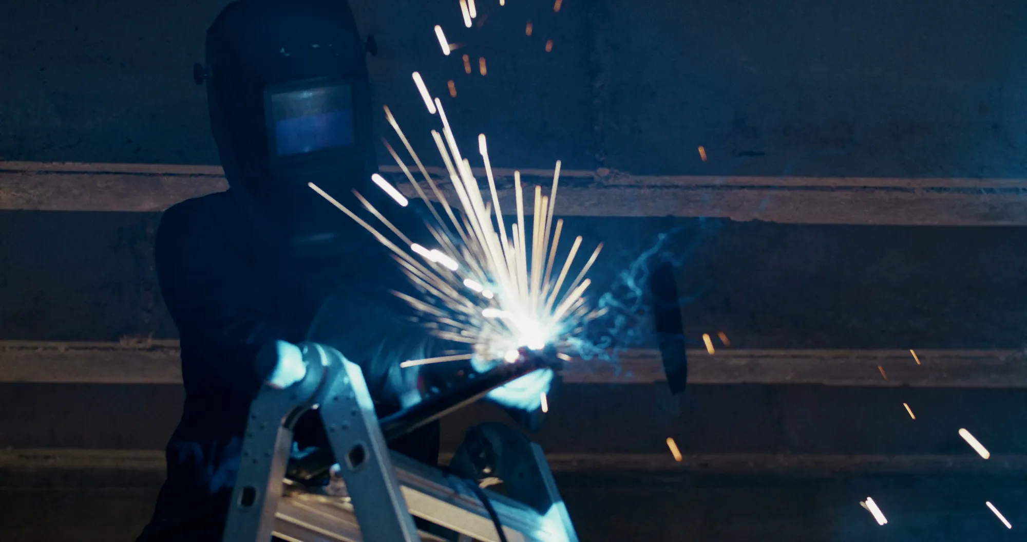 A welder in a dark auto-darkening helmet works at the top of a stepladder in a concrete interior, blue-white sparks bursting from the joint and lighting the visor.
