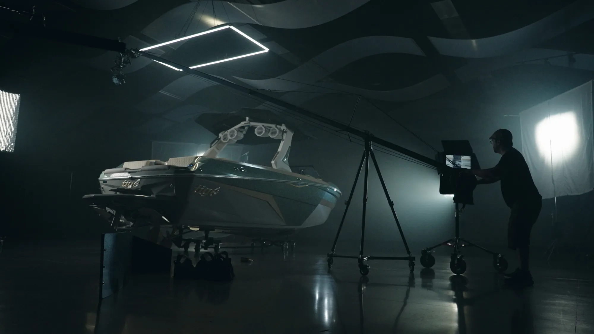 A blacked-out studio stage: a wake boat on low stands lit from the left by a large soft source, an angular LED tube hung overhead, a stand and flag mid-frame, and a crew member at a monitor cart on the right, all of it reflected in the polished floor.