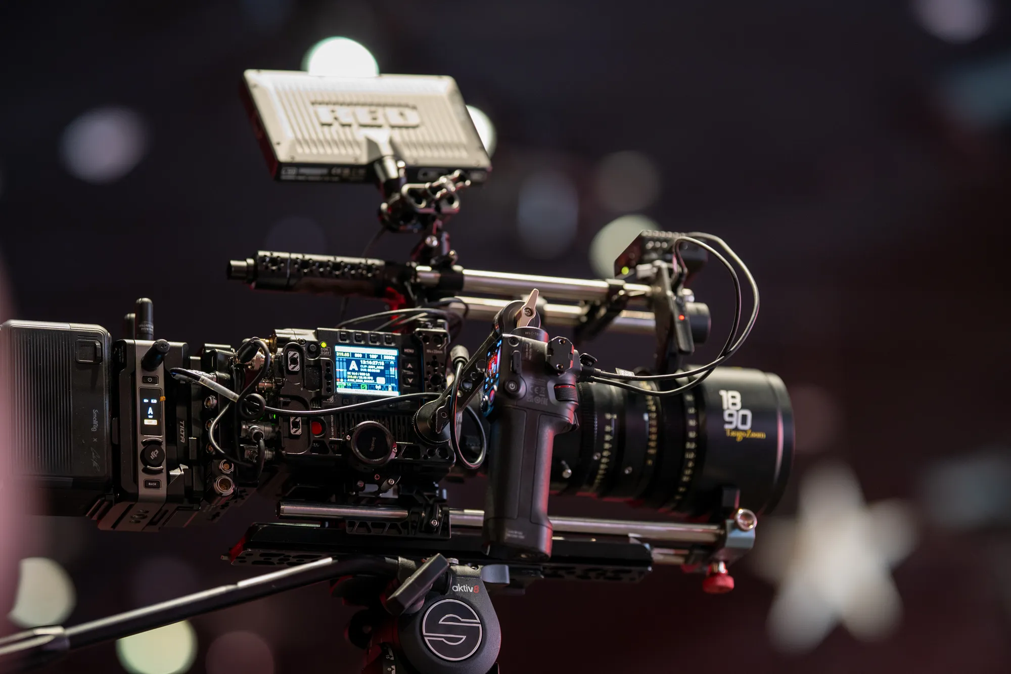 A cinema camera built up on a fluid head: monitor on top, cine zoom with marked focus and iris rings, side battery plate, cabling and a handle, against soft warm out-of-focus lights.