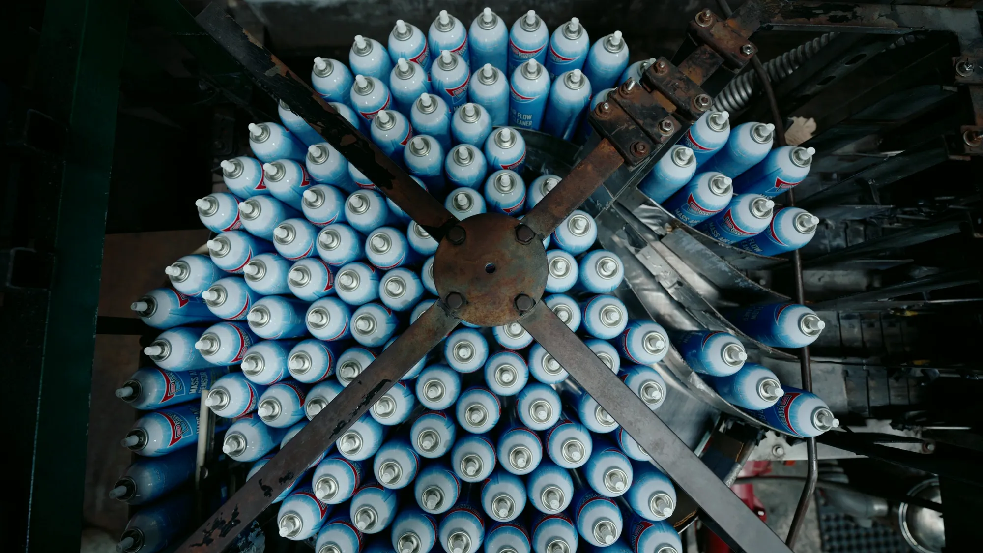 Straight down onto a rotary filling machine: pale blue aerosol cans packed in a dense circle around a steel cross, dark machinery around the edge of the frame.